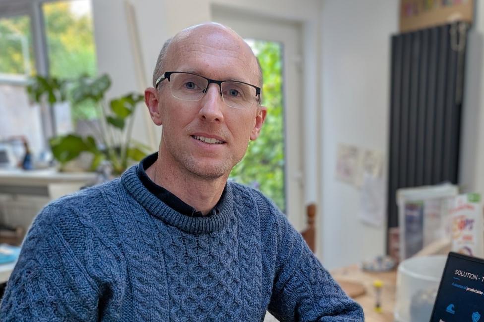 Portrait of a person wearing glasses and a blue knitted sweater, seated at a table in a bright home setting with plants and a laptop visible in the background.
