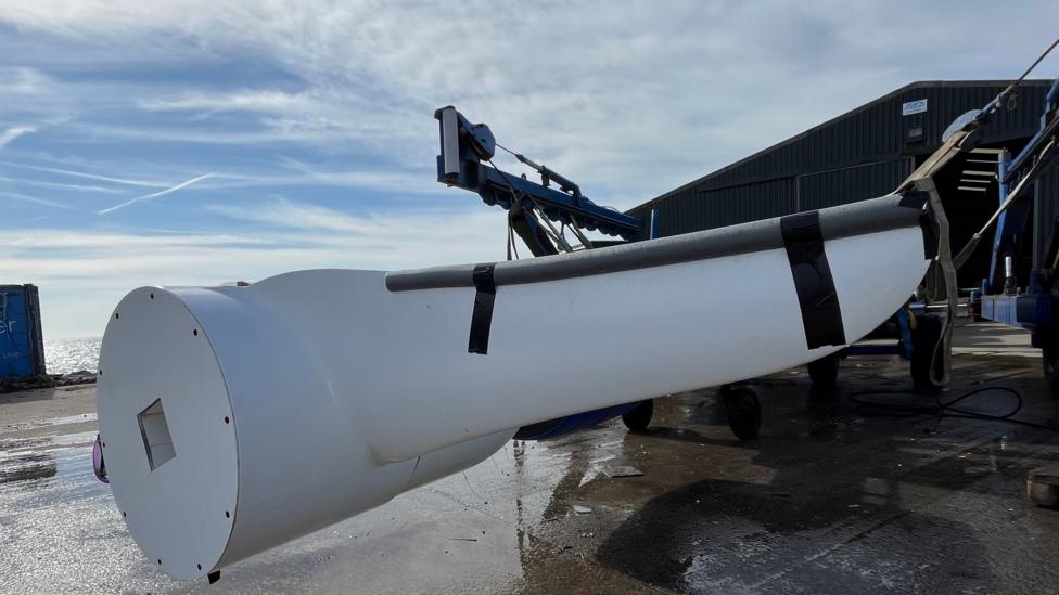 Large white marine turbine or underwater energy device being lifted by a crane outside a waterside industrial workshop, with boats and harbour facilities in the background.