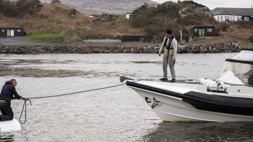 A small rigid inflatable boat in a harbour is connected by a rope to another vessel at left. One person stands on the bow of the boat while another handles the rope from the partially submerged vessel. Shoreline cabins, rocky embankments, and hills are visible in the background under an overcast sky.