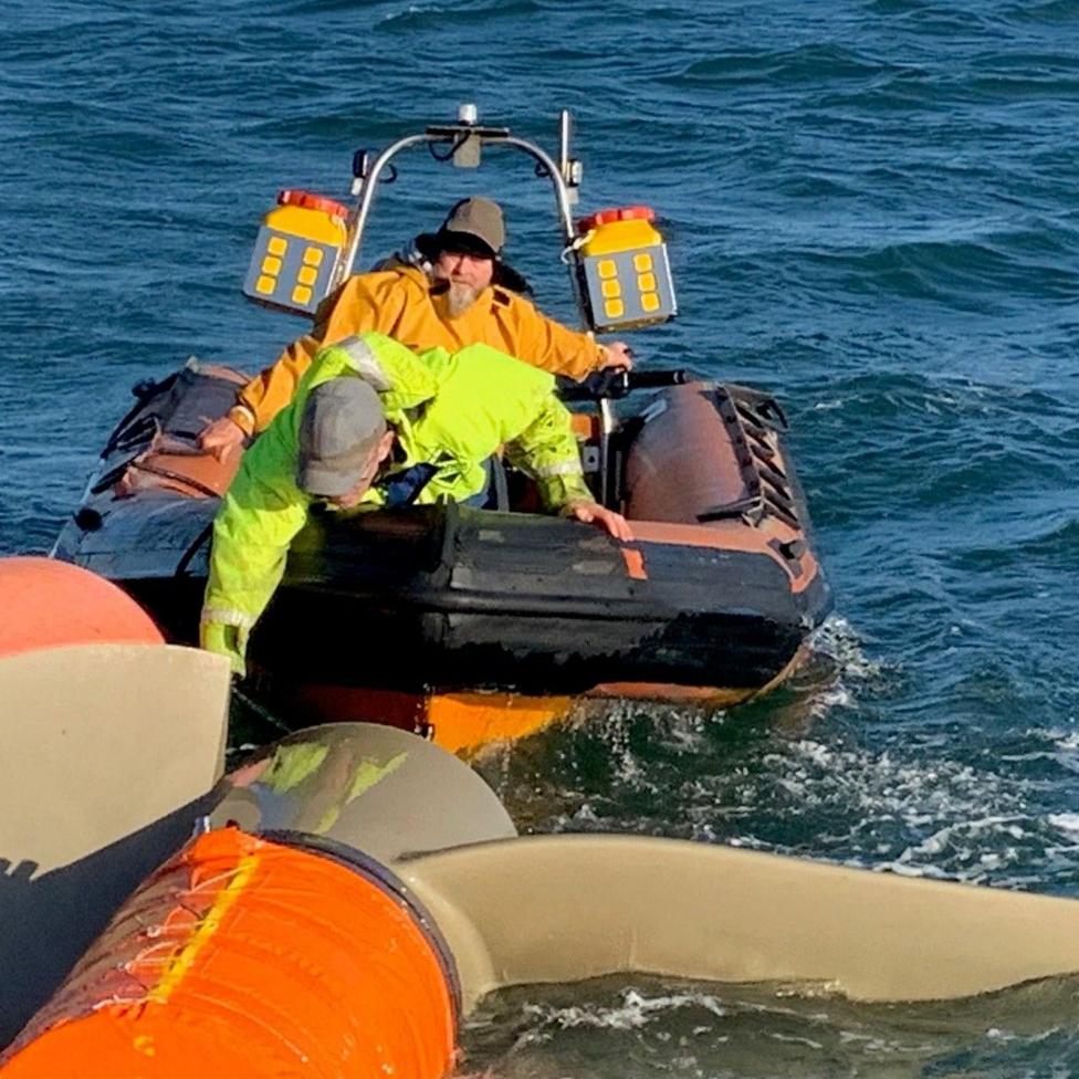 Two workers in a small inflatable workboat approach a floating tidal turbine structure at sea. One worker leans over the side of the boat toward the turbine component while the other operates the vessel. The scene shows offshore maintenance or service operations in choppy blue water under clear daylight conditions.