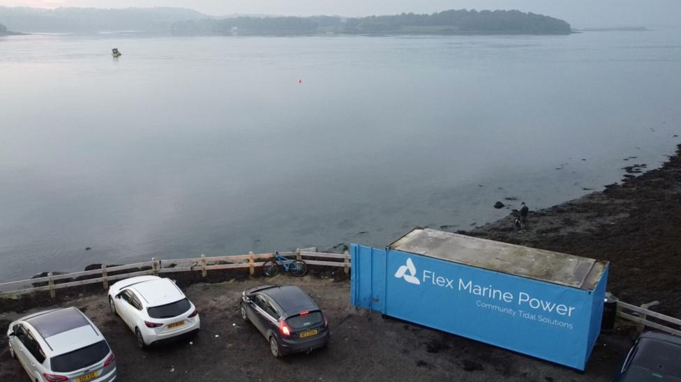 Aerial view of a coastal site with a blue shipping container branded “Flex Marine Power – Community Tidal Solutions” beside a small parking area with several cars. The container sits next to a rocky shoreline overlooking calm water, with a small vessel visible offshore and wooded land in the distance under hazy skies.