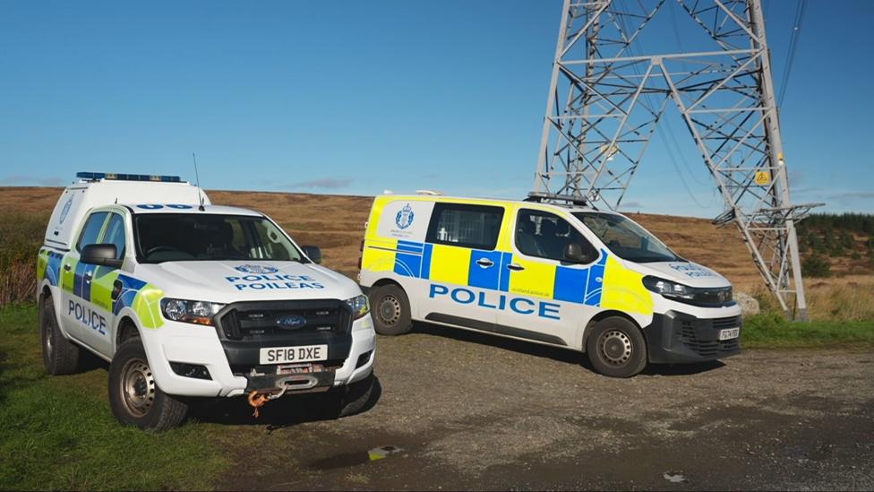 Police vehicles parked in a field by a pylon in daylight