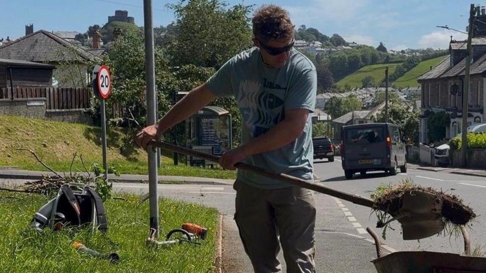 Man wearing sunglasses is about to drop a shovel full of soil and weeds and drop it into a wheelbarrow. He is wearing a blue t-shirt and khaki trousers. A strimmer and rucksack and other vegetation sits on a green area next to the pavement.