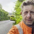 Man standing in the forefront of the photograph on a pavement with leaves from trees and bushes pushed up against him. A lorry is driving past to show the proximity of the overgrown pavement to the road.