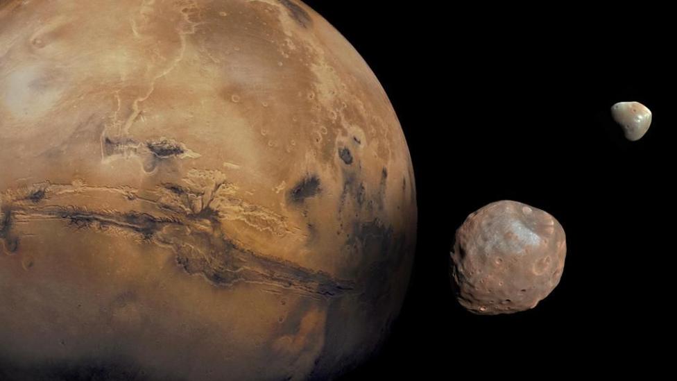 Mars fills most of the scene - it is orange and its surface is pitted with craters and canyons. Next to Mars, and against the blackness of space, there are its two small, irregularly shaped moons. In the foreground, the larger moon Phobos is pale red and covered in craters. In the distance, the smaller moon, Deimos, is grey and orange with an irregular shape.