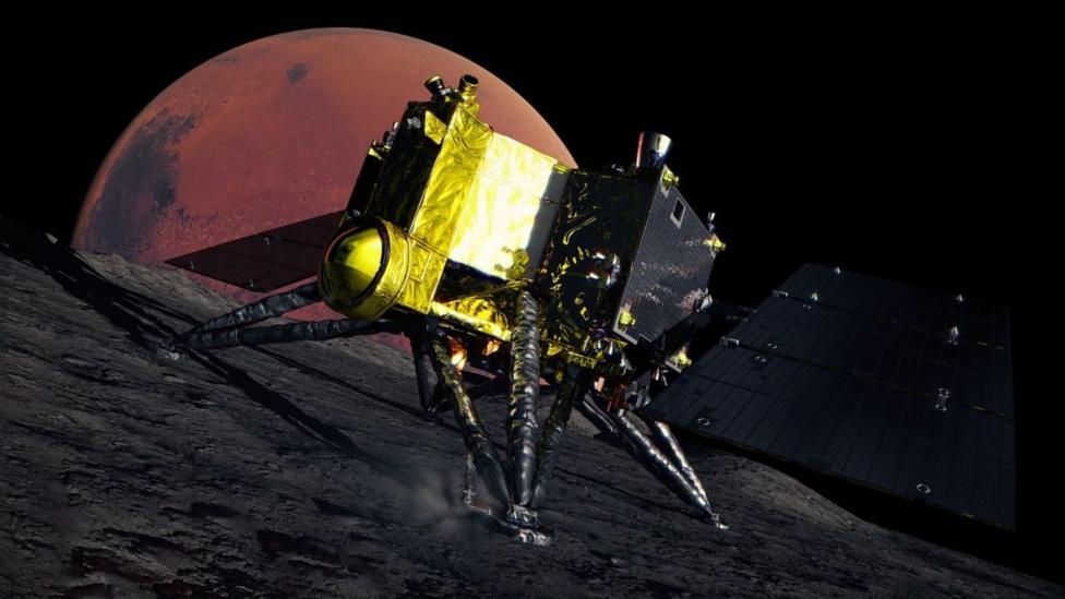 An artist's impression of the MMX spacecraft on the dark, rocky surface of Phobos. The spacecraft is gold and black and has four triangular legs and dark solar panels. The red disc of Mars is in the background.