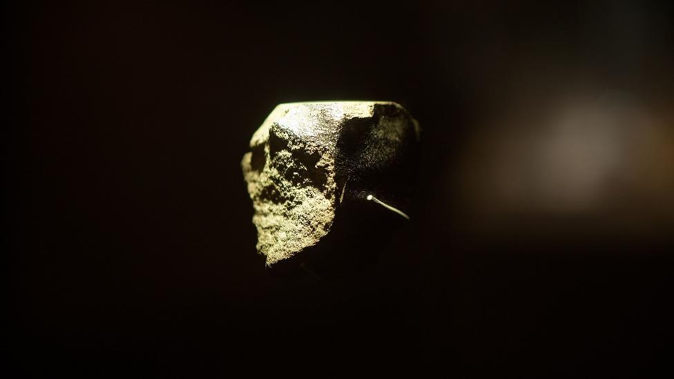 A close up image of a Martian meteorite which is on display at the Natural History Museum. It is small and irregularly shaped, it is lit from one side showing its rough texture. It is against a black backdrop of the display cabinet.