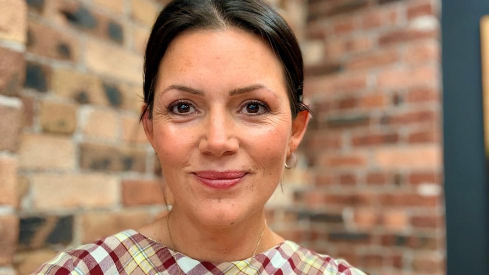 A woman with tied back brown hair and earings in a check top, standing in front of a bare brick wall in her office
