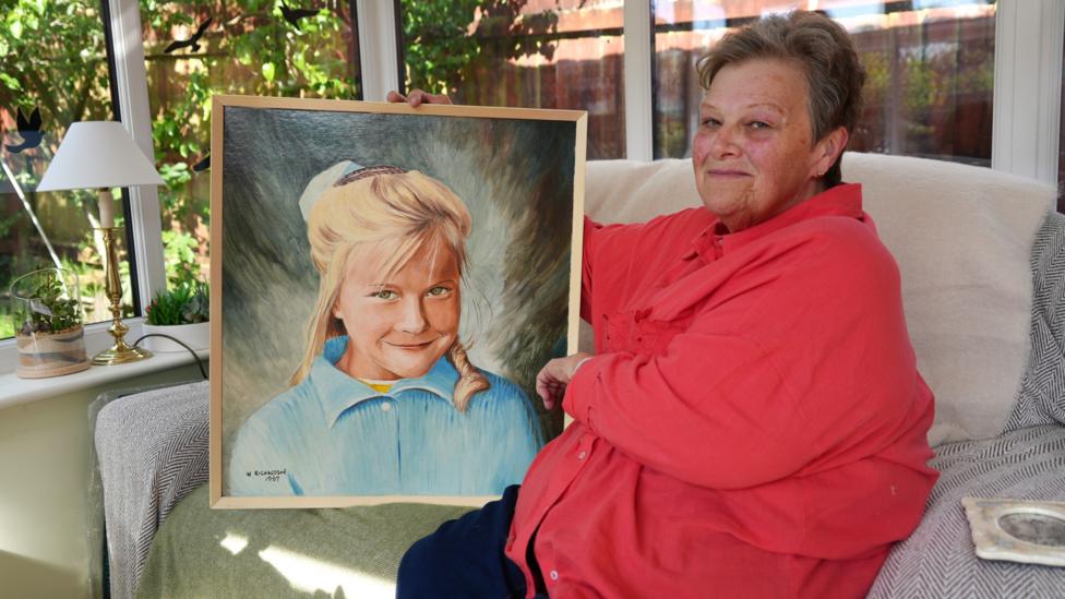 Jeanette Keane sits on her sofa wearing a red shirt, holding a portrait of her blonde-haired younger sister Edwina
