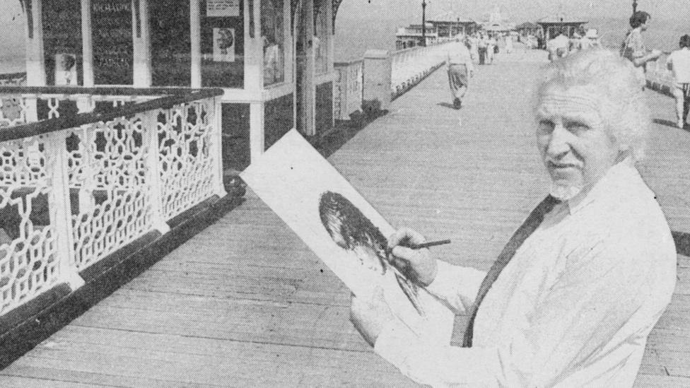 A black and white photo of a smartly-dressed sketch artist, holding a sketchpad and pencil on Llandudno Pier