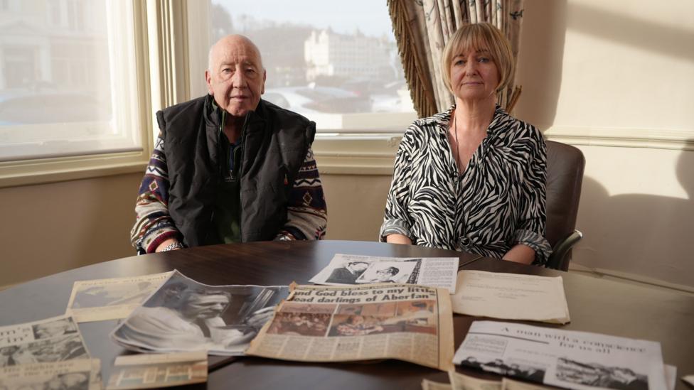 A man and woman sitting at a table that's covered in news paper cuttings and photographs