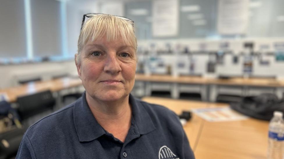 Karen Pyle is sitting at a classroom desk with the background slightly out of focus. Behind her there are desks and chairs and a plastic water bottle on the table to her right. She has blonde hair, tied back and with a fringe. She has a pair of glasses perched on her head and she is wearing a dark blue polo shirt with the white edge of a the Newcastle College bridge logo just showing.