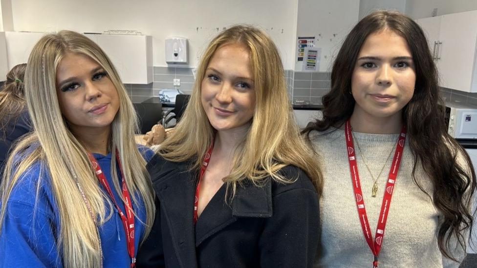 Three teenage girls are standing. The first going left to right has long blonde hair and is wearing a cobalt blue jumper. In the middle she also has long blonde hair and a navy jacket, on the right is Charlotte and she is wearing an oatmeal knitted round neck jumper and has long dark hair. All three are wearing their college lanyards which have red straps. They're standing in a room in the college and behind them is a kitten with pale grey metro tiles and white cabinets. 