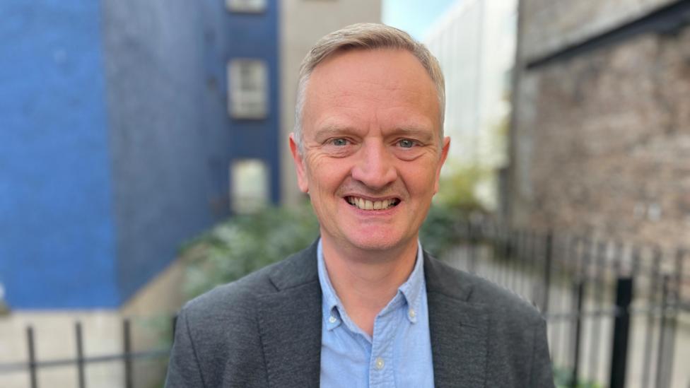 Paul White. He is wearing a blue shirt and grey blazer. He stands outside with a blue building behind him.