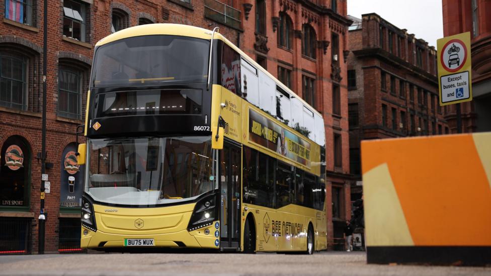 Yellow Bee Network electric bus in Manchester. Red sandstone buildings stand on either side of the street.