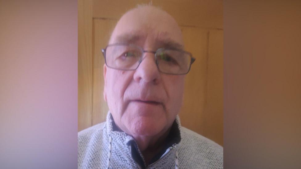 An older bald man who is wearing glasses and a grey woollen top with a high collar.
