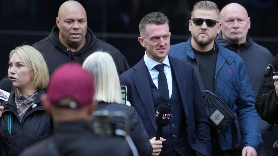 Tommy Robinson (centre) arrives at Southwark Crown Court. He holds a black microphone with the letters UTK on the cover, coloured in the British flag colours. A group of others accompany him. He wears a navy blue suit with a white shirt and blue tie.