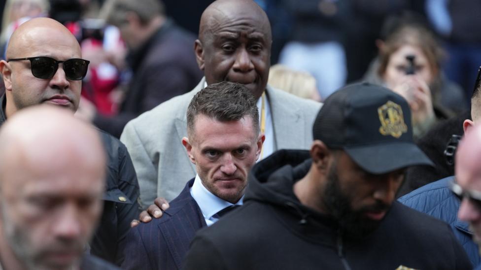 Tommy Robinson is in the midst of a group of people who appear to be security or police staff. One man standing behind him rests his hand on Robinson's shoulder.