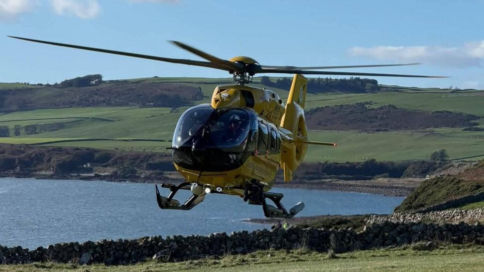 A yellow helimed helicopter is a few feet above the ground on the Solway coast with hills, cliffs and the sea behind it