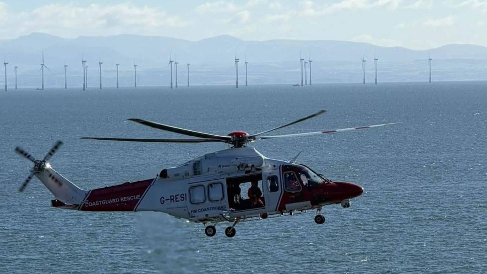 A coastguard helicopter over the waters of the Solway with wind turbines in the background and the hills of Cumbria