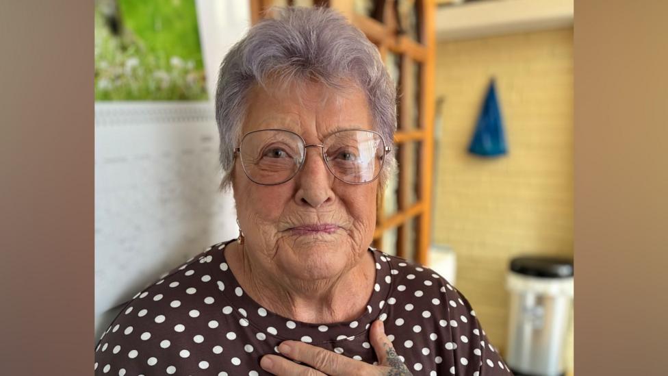 Portrait of an older person with short grey hair and round glasses, wearing a brown top with white polka dots, seated indoors with one hand resting on their chest. Shelving, a wall calendar and household items are visible in the softly blurred background.