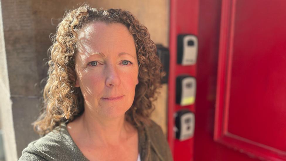 Katie Beale is standing in front of her red door, there are three key boxes behind her. She has brown curly hair and is looking directly at the camera.