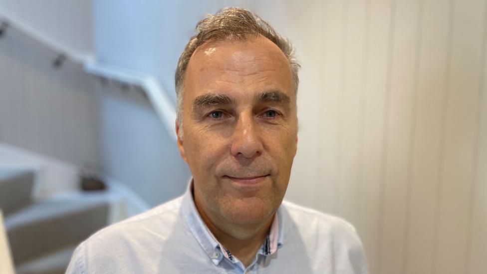 Paul Phillips is in his fifties and is wearing a light blue shirt. He is standing in front of a staircase