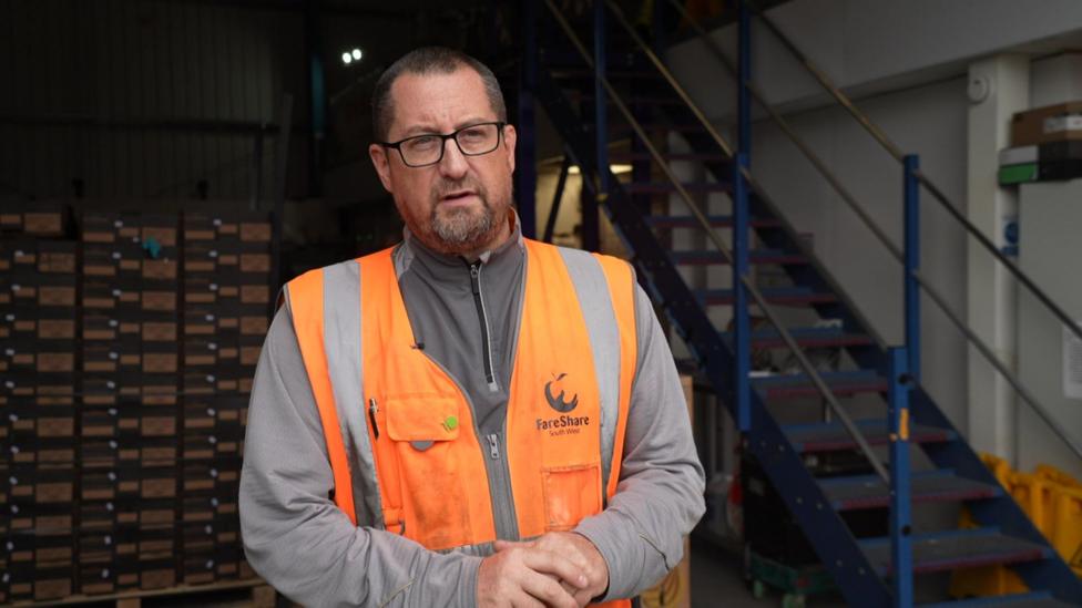 Ryan Jackson, Plymouth Hub Manager for FareShare South West, stands inside the charity's distribution hub wearing a yellow high visibility tabard and black-framed glasses.