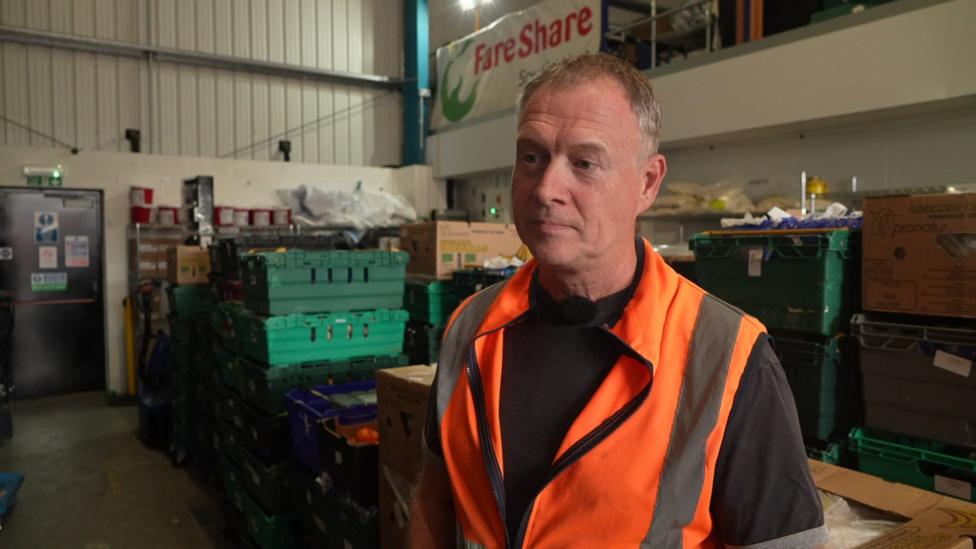 Jamie Henshaw, a volunteer with FareShare South West, stands inside the charity's distribution hub. He is wearing a black T-shirt and an orange high visibility tabard, with a large FareShare sign behind him.