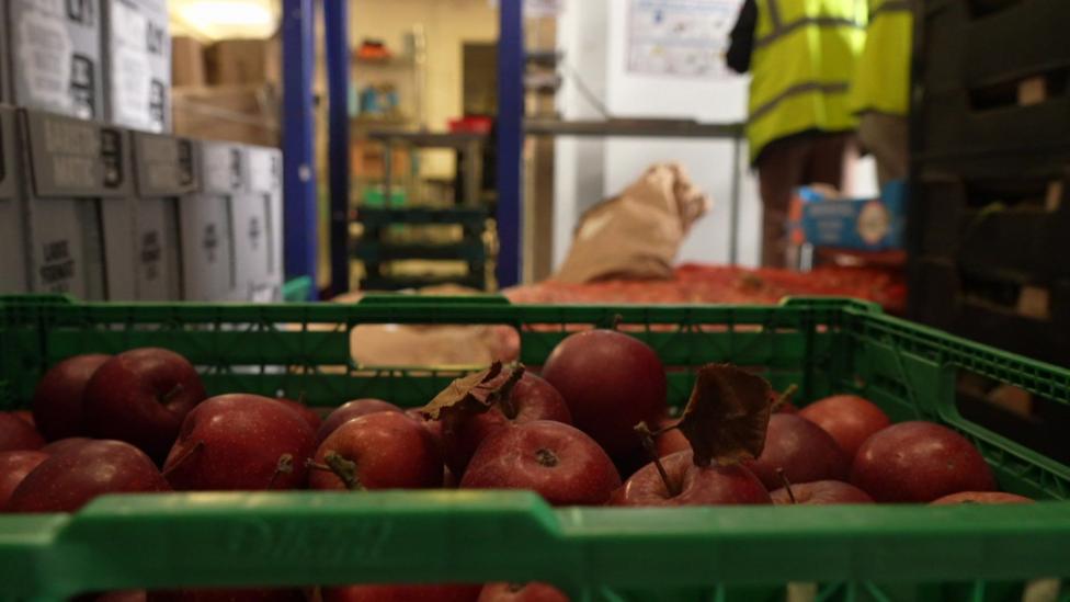 Close-up of a green plastic crate filled with red apples at FareShare South West's distribution hub in Plymouth. Two volunteers can be seen out of focus in the background.