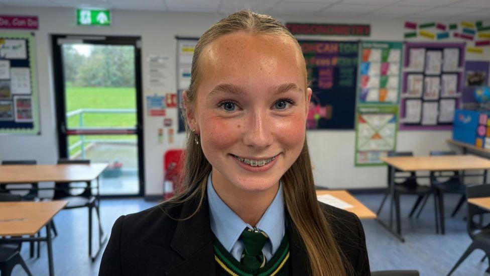 A female pupil in a school uniform stands in the centre of a brightly lit classroom, facing the camera. The classroom contains desks and chairs, colourful educational displays on the walls, a door leading outside in the background. The student has long hair tied back and is wearing a blazer, shirt, tie and green jumper.