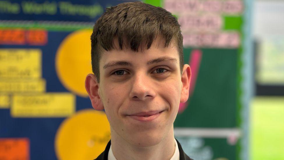 Head and shoulders portrait of a male student with brown hair in a classroom, facing the camera. The student is wearing a dark school blazer and light-coloured shirt. Behind the student are blurred classroom displays featuring colourful educational posters.