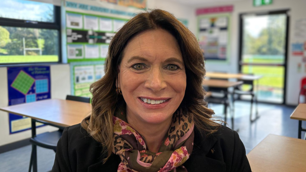 A woman with shoulder length brown hair sits at a desk in a classroom, facing the camera. She is wearing a dark jacket and a pink and brown patterned scarf. The classroom contains tables and chairs, educational posters and displays on the walls, and a glass door showing a grassy outdoor area.
