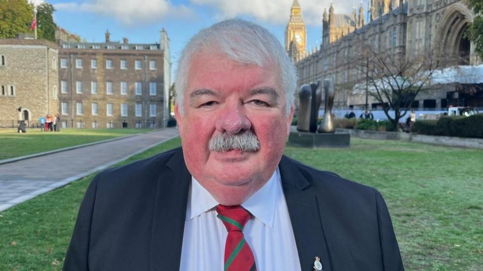 A man with short grey hair and a short grey moustache wears a black suit, white shirt and red tie. He stands in a green-grass pavilion in front of light brown buildings. 