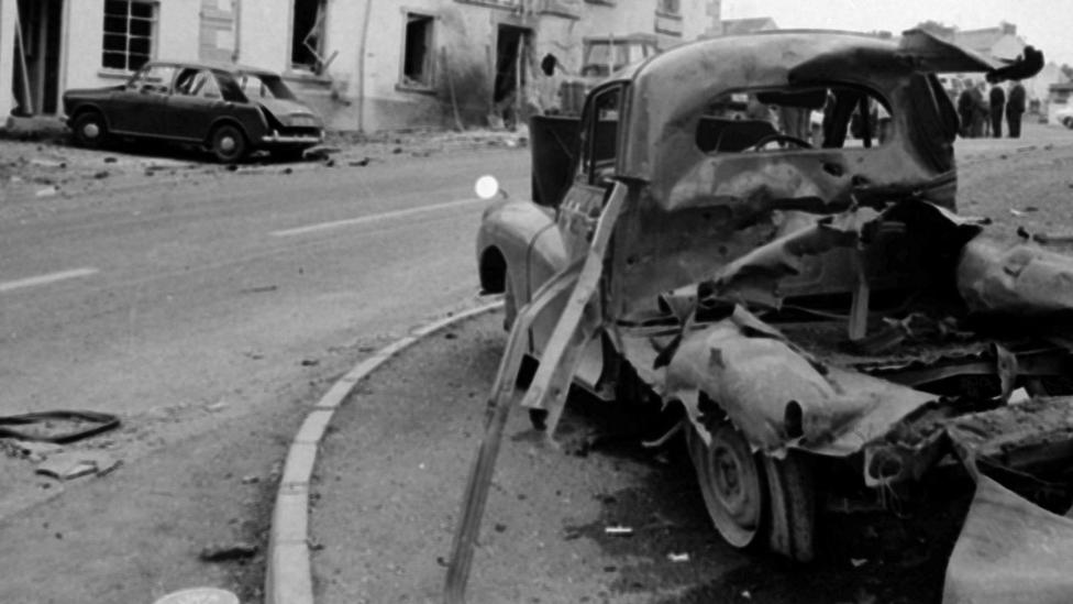 A black and white photo showing the aftermath of bombs. There are damaged cars and a damaged building in the photo.