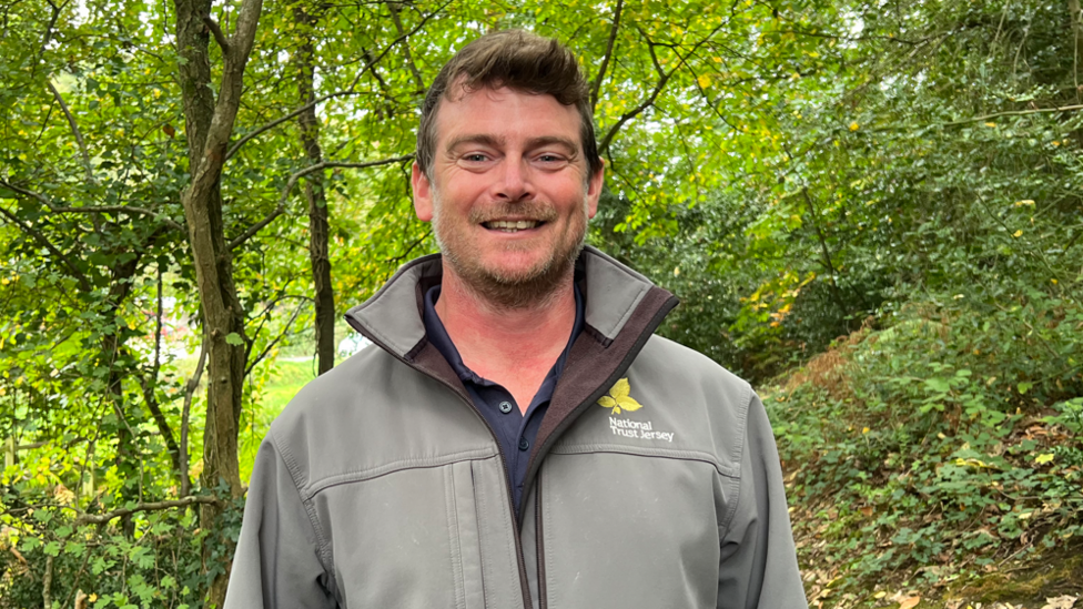 Henry Glynn is pictured smiling in a woodland. He is wearing a green National Trust coat. He has short brown hair and stubble.