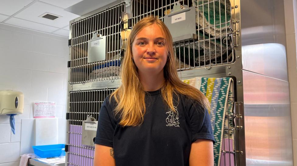 Nina Morgan pictured in the JSPCA office. There are cages visible behind her. She is wearing a navy JSPCA tshirts. She has long blonde hair and is smiling.