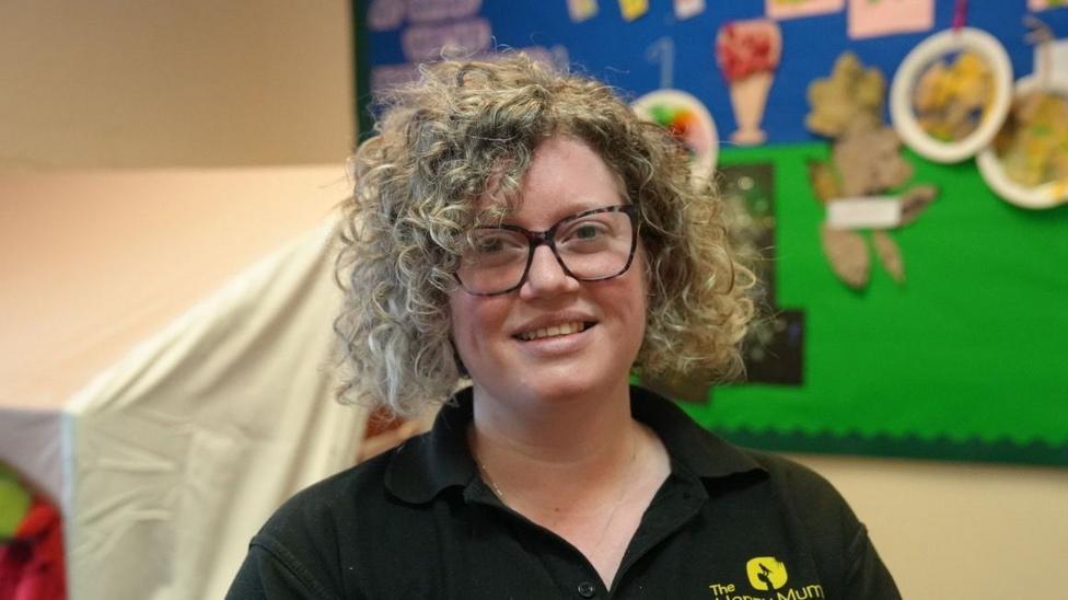 Lucinda Bray has blonde curly bobbed hair. She is wearing glasses and smiling towards the camera. She is wearing a black polo top with a Happy Mums logo on the front in yellow.