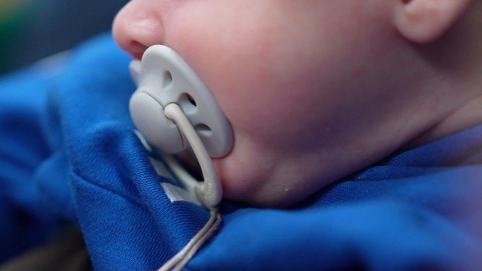 This is a picture of an anonymous baby, who is dressed in blue and has a pacifier.