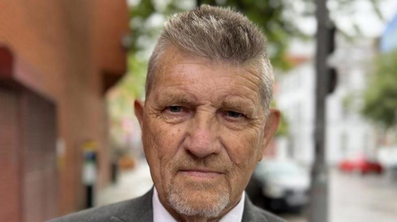 Shows a man with a beard. He wearing a grey jacket with a white shirt and blue striped tie.