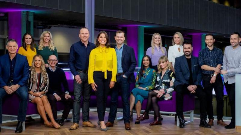 A crowd of TV presenters, both men and women, sitting on and gathered around a purple sofa.