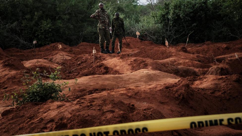 Two men in military uniform stand on red earth where several large holes have been dug up. Behind them tress can be seen. In the foreground is a yellow banner which reads: Crime scene do not cross