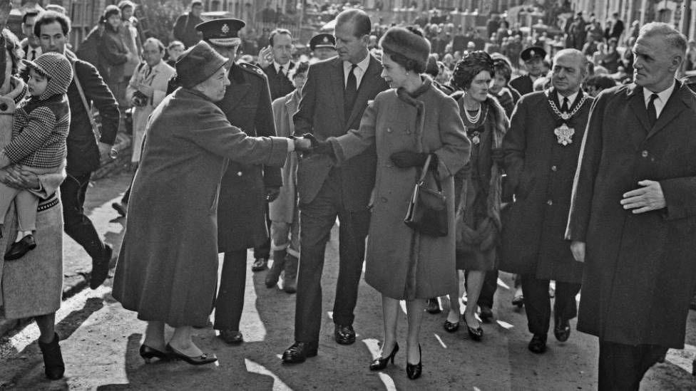 A black and white image of Queen Elizabeth meeting people in Aberfan. She shakes a woman's hand, with crowds of people around her. She wears a fur hat, and fur-lined coat.