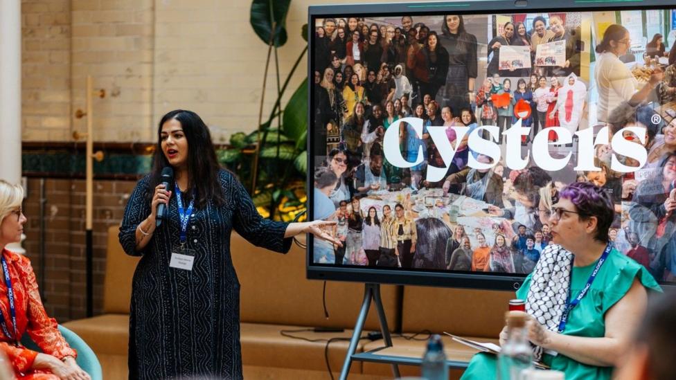 A woman speaks into a microphone during a panel discussion beside a large display screen showing photographs from community events. The word Cysters can be seen behind her.