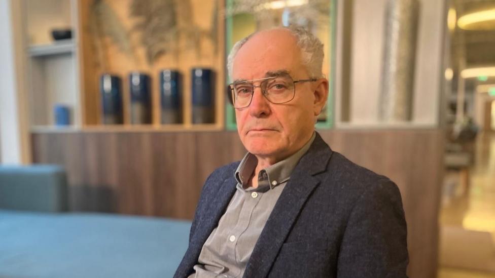 Clive Ruggles, a man with glasses and grey balding hair looks at the camera with a serious expression. He wears a dark textured blazer and grey shirt. He sits in a wood-panelled room with blue upholstery.