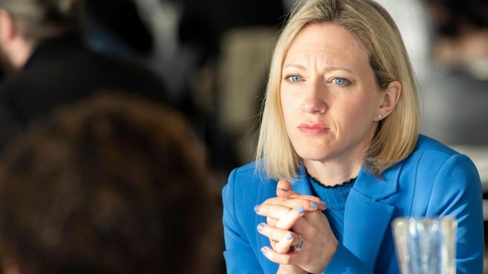 Claire Waxman, a blonde-haired woman with blue eyes wearing a blue jacket and blue ribbed top, clasps her hands together. She is wearing blue nail varnish and has a serious expression. There are people out-of-focus in the background.