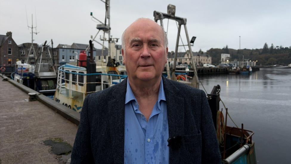 A man wearing a blue shirt and dark blue jacket standing on a pier next to boats in a harbour with a grey blue sky