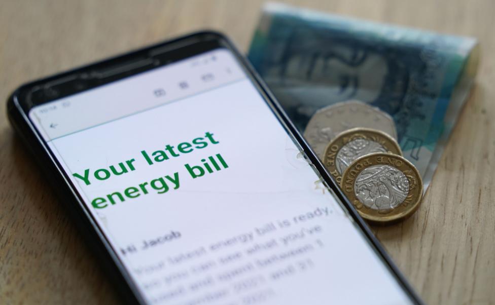 Photo of an online energy bill on the screen of a mobile phone next to a folded five pound note, two pound coins and a 50p coin