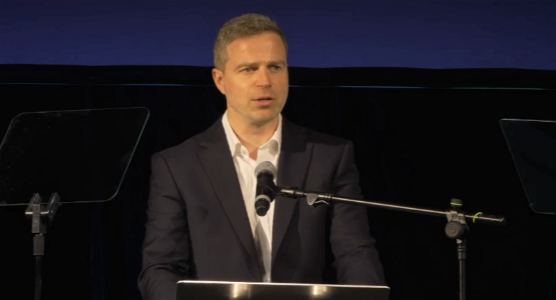 Duncan Parish, wearing a black jacket and white shirt, speaks at a podium with a microphone in front of him.