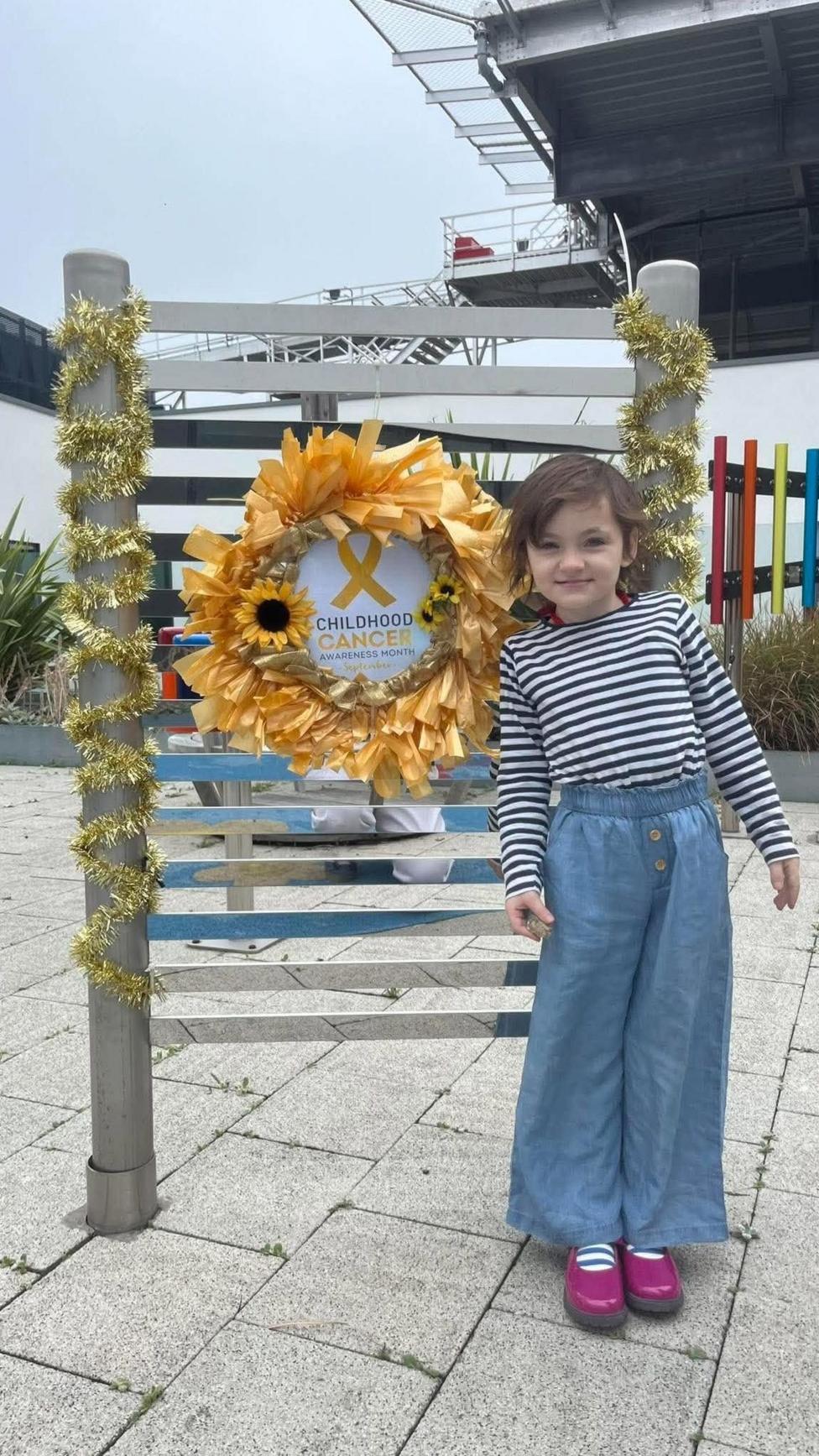 Flora stands beside a decorated outdoor display featuring a large gold wreath with sunflower accents and a yellow ribbon. The sign in the centre reads “Childhood Cancer Awareness Month.”